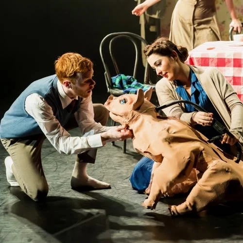 Two performers kneel beside a large pig puppet on stage, with a red-and-white checked table behind them.