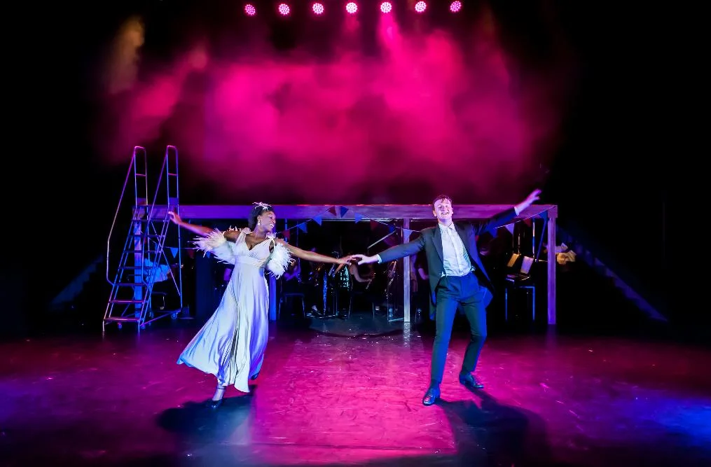 Two performers in a pale flowing dress and dark suit dance hand in hand beneath pink stage lights, in front of a raised band platform.