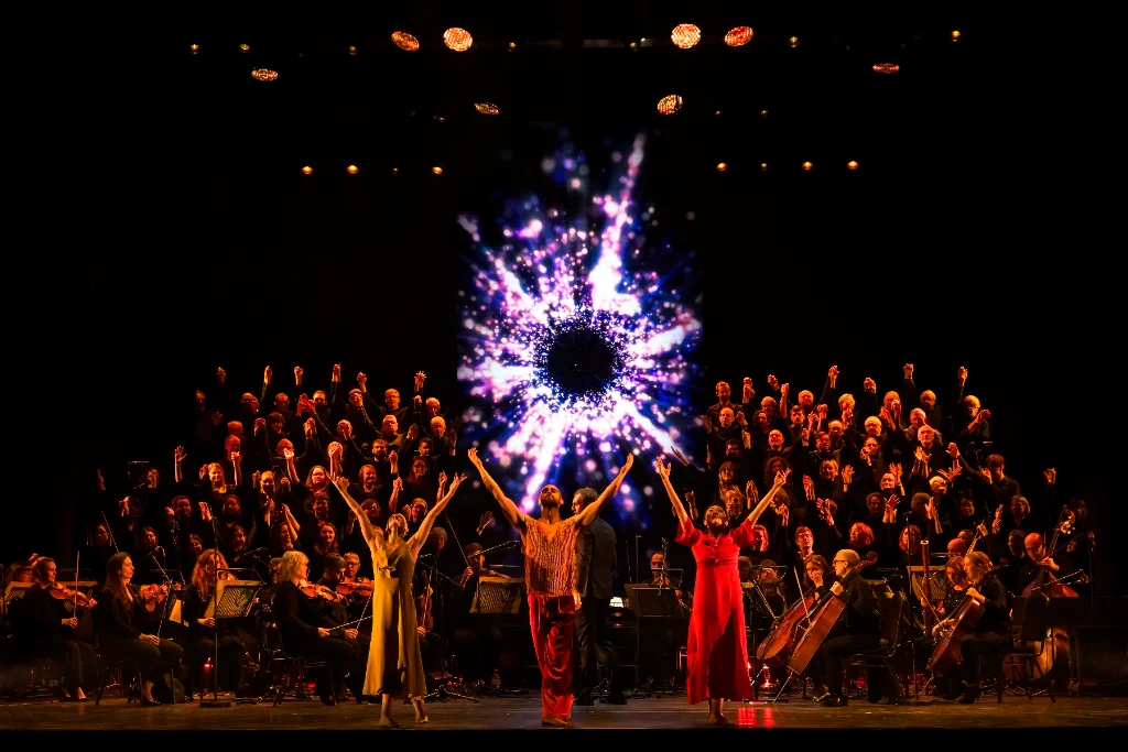 Three dancers raise their arms before an orchestra and choir, with a bright purple circular projection at the centre of the stage.