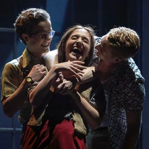Three performers embrace and laugh together under blue stage lighting.