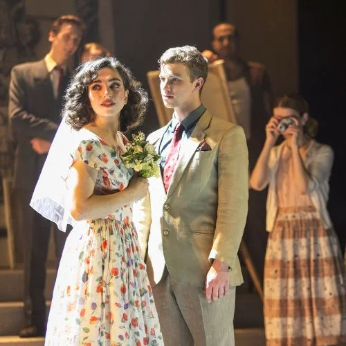 A woman in a floral wedding dress and veil stands beside a man in a light-coloured suit, with other performers behind them.